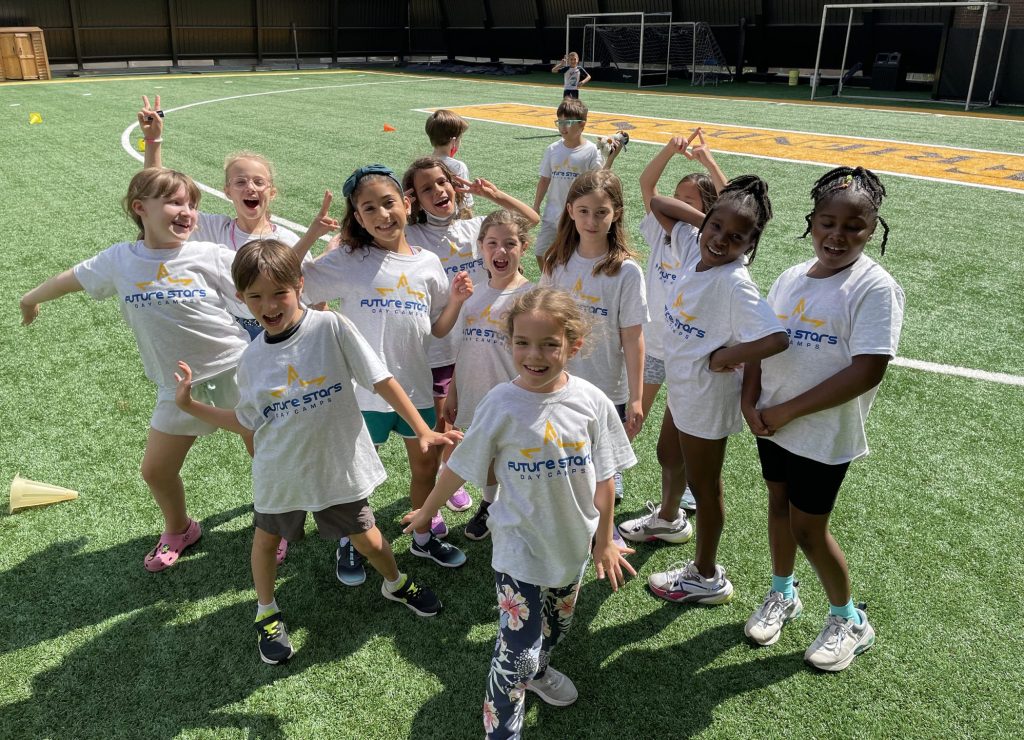 A photo of a group of young girls as the focal point, posing and smiling on a turf field on a sunny day at day camp