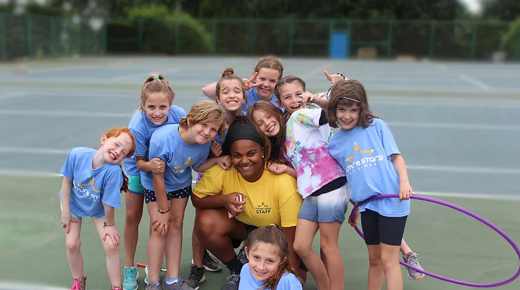 a group of young girls surrounding and posing with a young female camp counselors on an outdoor sports court